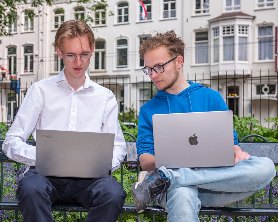 Twee middelbare scholieren werken buiten op een bank met laptops in Amsterdam.